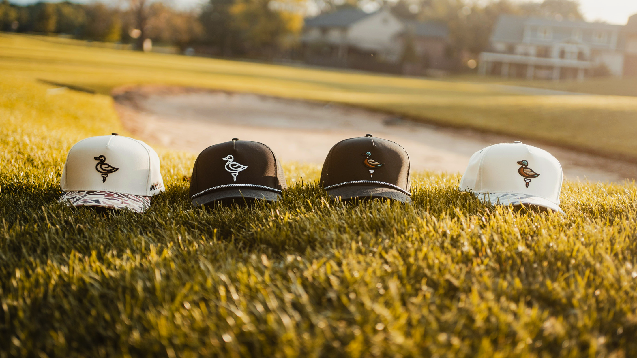 Four golf hats, with logos of a duck on a tee, on golf course and trees and a sand trap in the background.