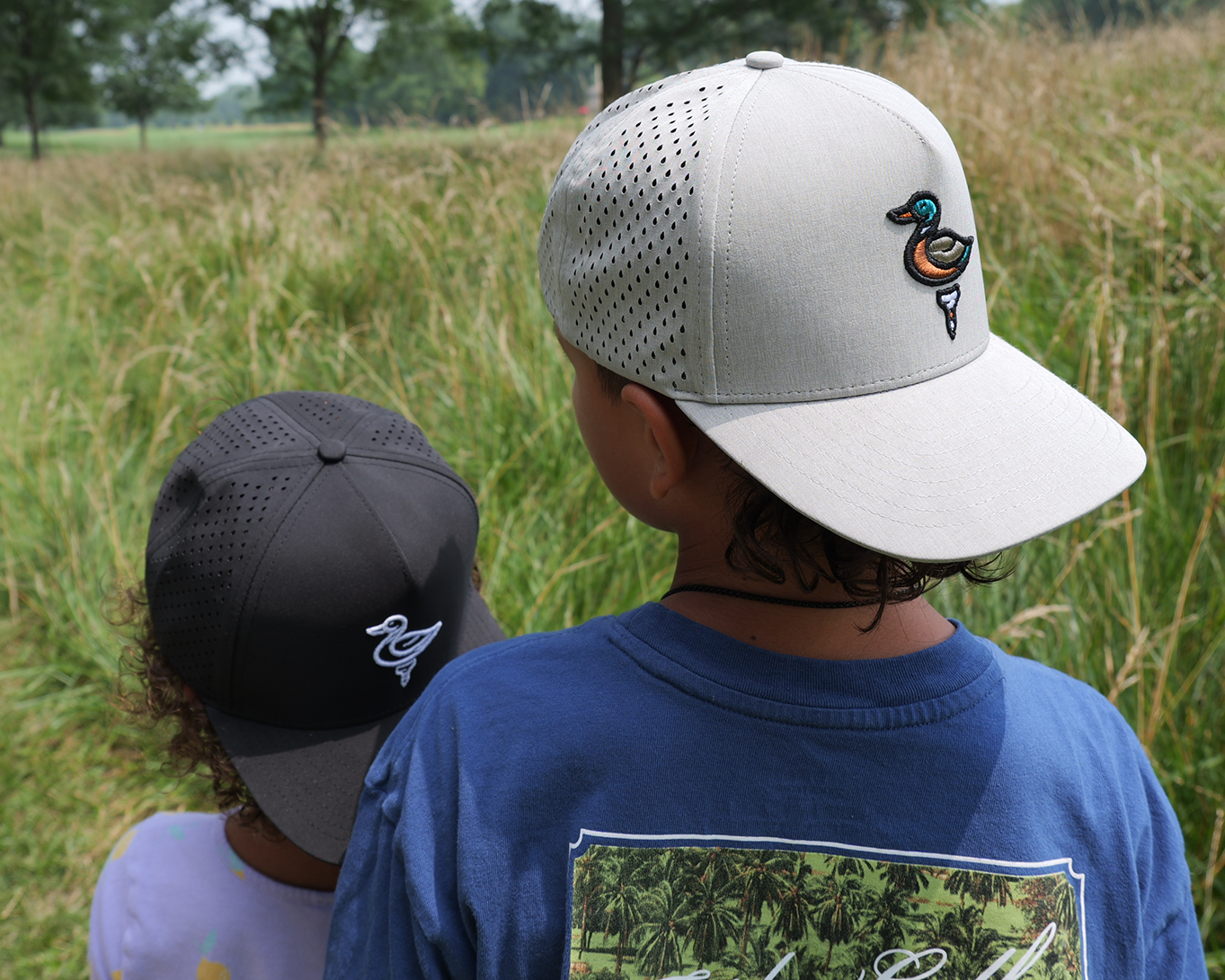 Two children seen from behind in a grassy outdoor field, each wearing a mesh baseball cap. The child in front wears a light gray cap with a small duck logo, and the child behind wears a dark gray cap with a white logo.