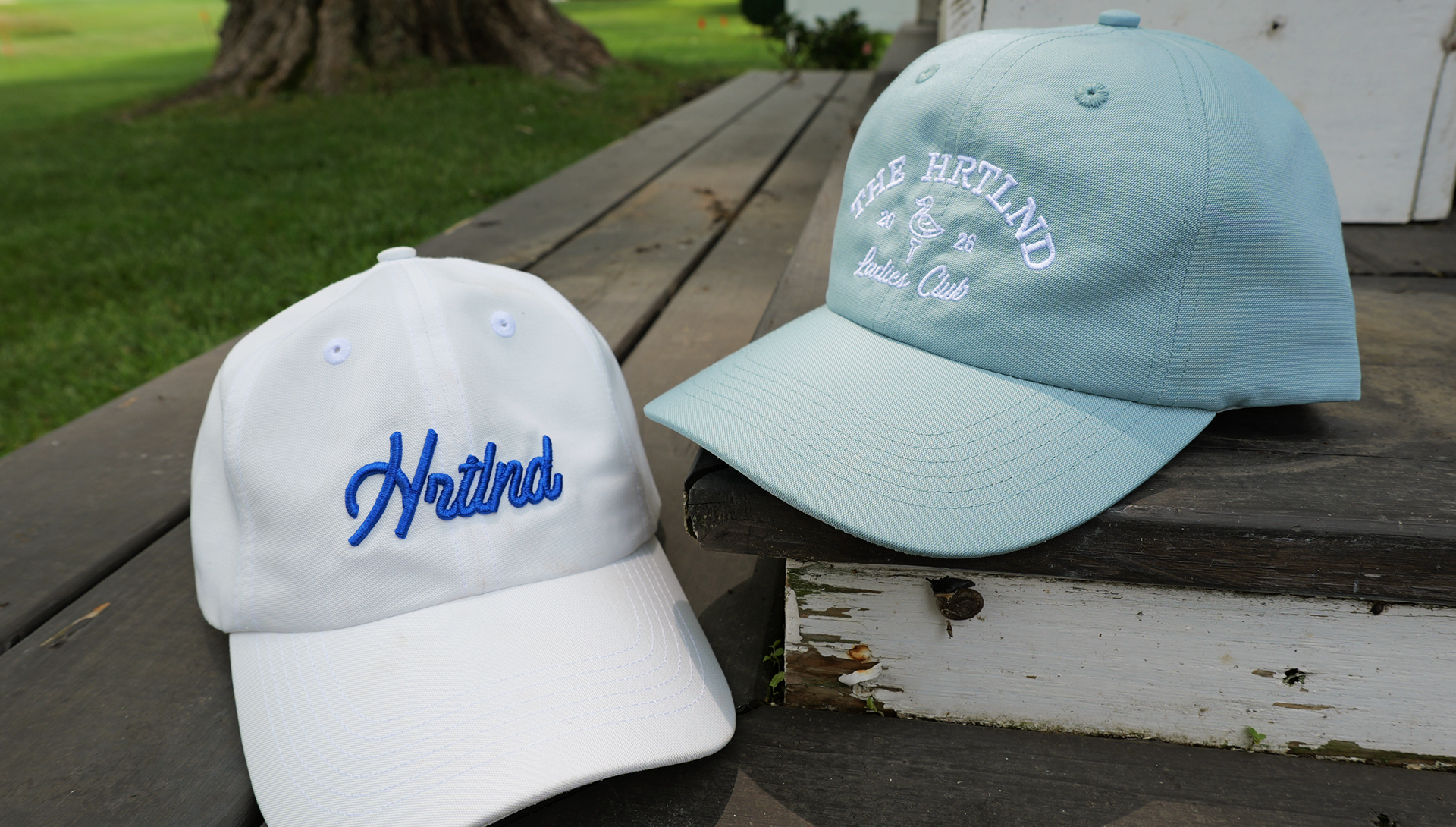Two baseball caps displayed outdoors on a weathered wooden porch. The cap on the left is white with blue script embroidery reading 'Hrtlnd'.