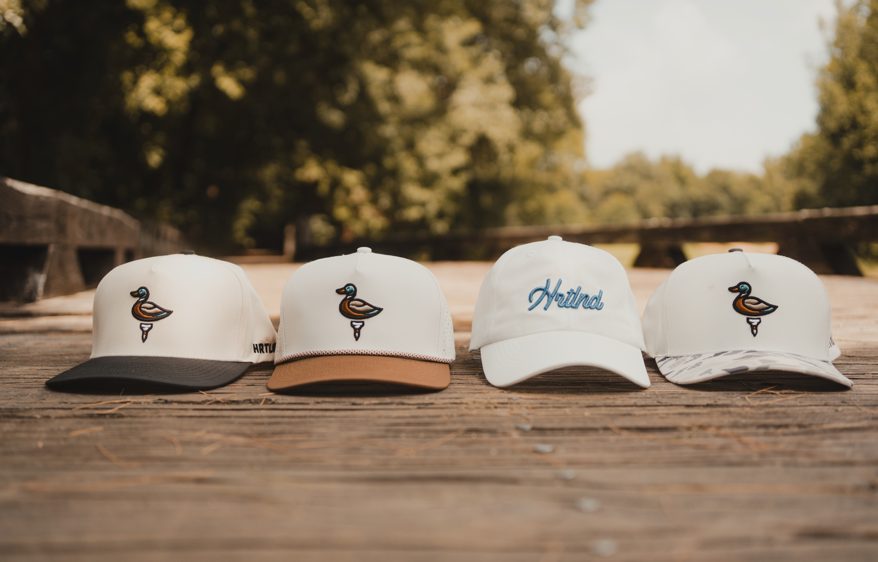 Four white snapback caps lined up on a wooden deck outdoors, each featuring a small embroidered duck logo and different brade colors, with a blurred forest background.